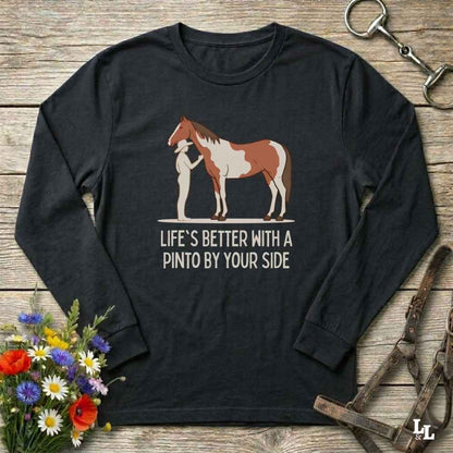 A black long-sleeve t-shirt with a graphic of a Pinto horse and a person, accompanied by the text 'LIFE'S BETTER WITH A PINTO BY SIDE'. The shirt is displayed flat on a rustic wooden background with decorative props including a bouquet of wildflowers, a horse bridle, and a metal snap hook.