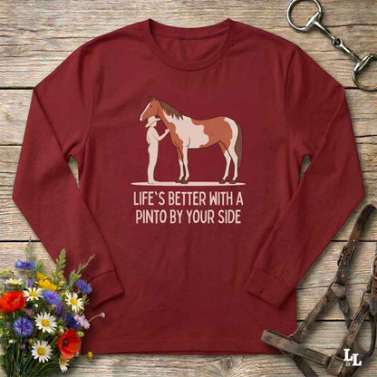 A long-sleeve maroon crewneck t-shirt laid flat on a rustic wooden surface, featuring a graphic of a person and a Pinto horse with the text 'LIFE'S BETTER WITH A PINTO BY YOUR SIDE'. The shirt is styled with equestrian and floral props.