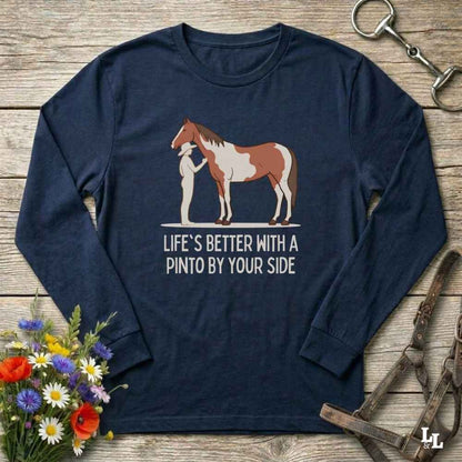 A navy blue long-sleeve t-shirt with a graphic of a Pinto horse and the text 'LIFE'S BETTER WITH A PINTO BY YOUR SIDE', displayed in a flat-lay arrangement on a rustic wooden background with a horse bridle, flowers, and a horse bit.