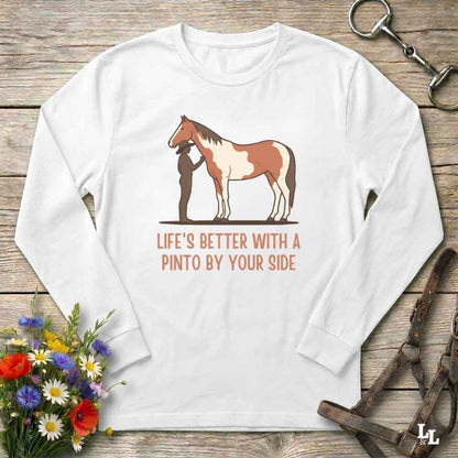 A white long-sleeve crewneck t-shirt with a graphic of a Pinto horse and a person, accompanied by the text 'LIFE'S BETTER WITH A PINTO BY YOUR SIDE' in orange. The shirt is displayed flat on a rustic wooden background with equestrian accessories and a small bouquet of wildflowers.