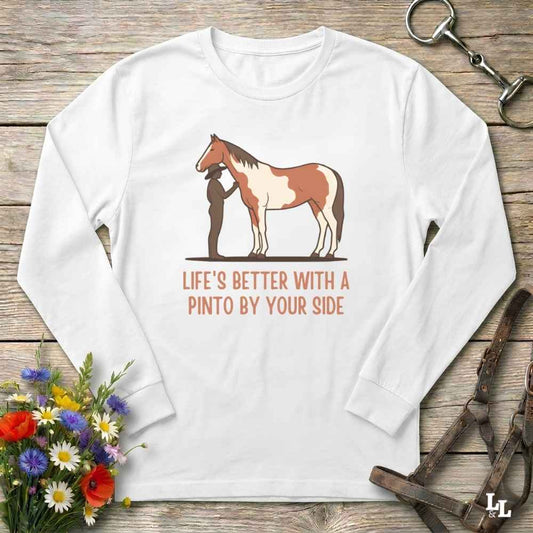 A white long-sleeve crewneck t-shirt with a graphic of a Pinto horse and a person, accompanied by the text 'LIFE'S BETTER WITH A PINTO BY YOUR SIDE' in orange. The shirt is displayed flat on a rustic wooden background with equestrian accessories and a small bouquet of wildflowers.