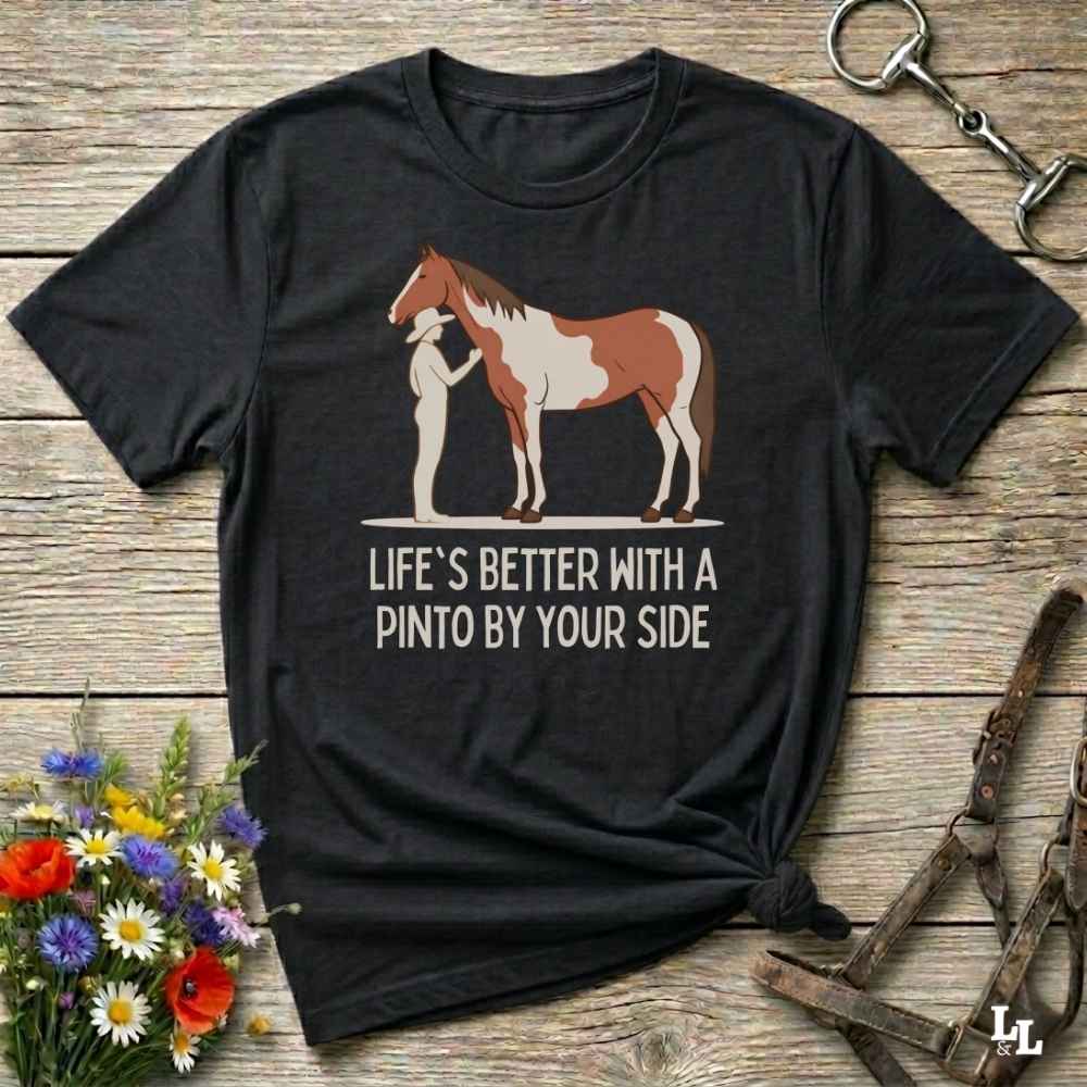 A black short-sleeve graphic T-shirt laid flat on a rustic wooden surface, featuring an illustration of a person standing beside a brown-and-white pinto horse with the text 'LIFE'S BETTER WITH A PINTO BY YOUR SIDE.' Decorative flowers and horse tack are placed around the shirt.