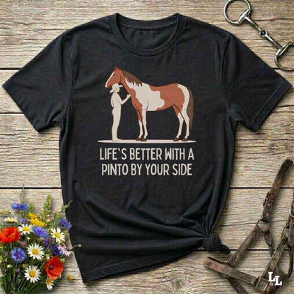 A black short-sleeve graphic T-shirt laid flat on a rustic wooden surface, featuring an illustration of a person standing beside a brown-and-white pinto horse with the text 'LIFE'S BETTER WITH A PINTO BY YOUR SIDE.' Decorative flowers and horse tack are placed around the shirt.
