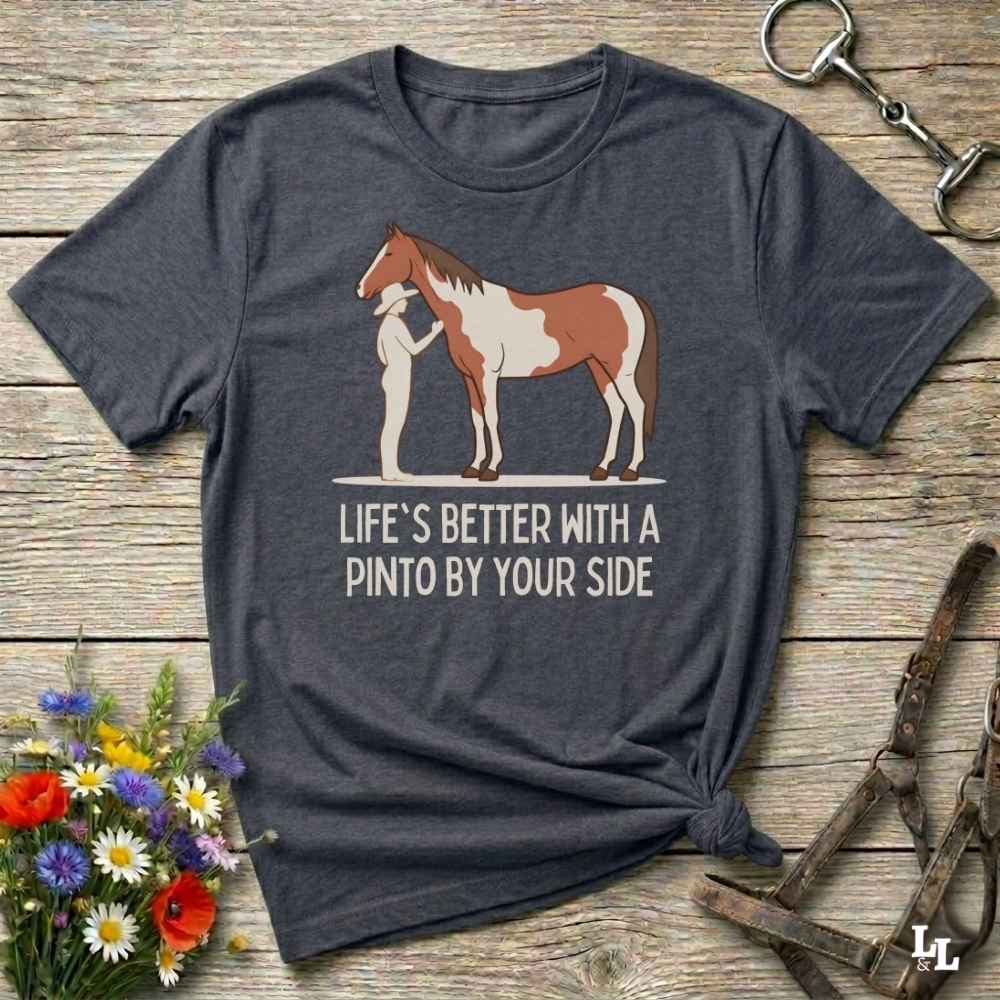 A dark charcoal gray t-shirt with a graphic of a Pinto horse and the text 'LIFE'S BETTER WITH A PINTO BY YOUR SIDE', displayed on a rustic wooden background with equestrian accessories and flowers.