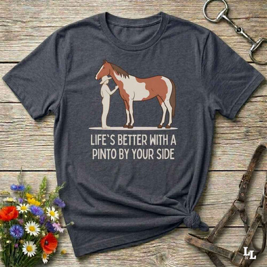 A dark charcoal gray t-shirt with a graphic of a Pinto horse and the text 'LIFE'S BETTER WITH A PINTO BY YOUR SIDE', displayed on a rustic wooden background with equestrian accessories and flowers.