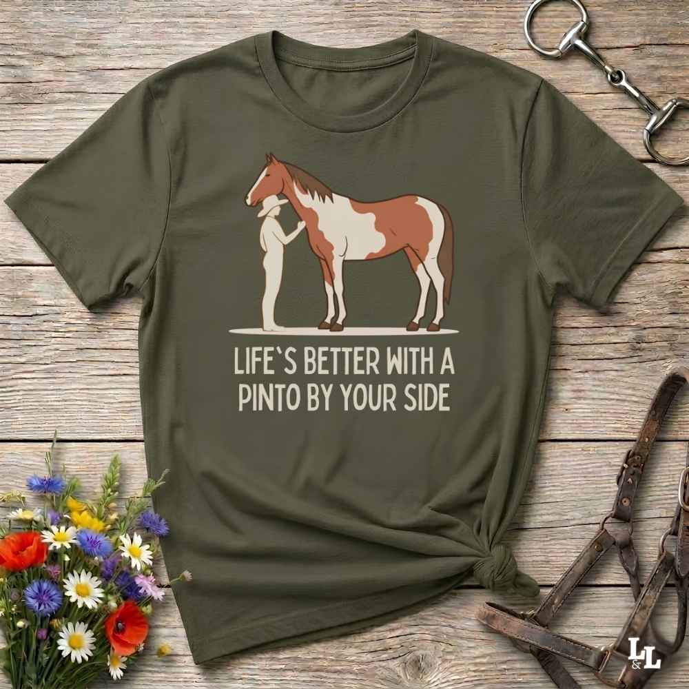 An olive green short-sleeve t-shirt with a graphic of a Pinto horse and a person, accompanied by the text 'LIFE'S BETTER WITH A PINTO BY YOUR SIDE'. The shirt is styled in a flat-lay on a rustic wooden background with a horse bridle, a horse bit, and a small bouquet of wildflowers.