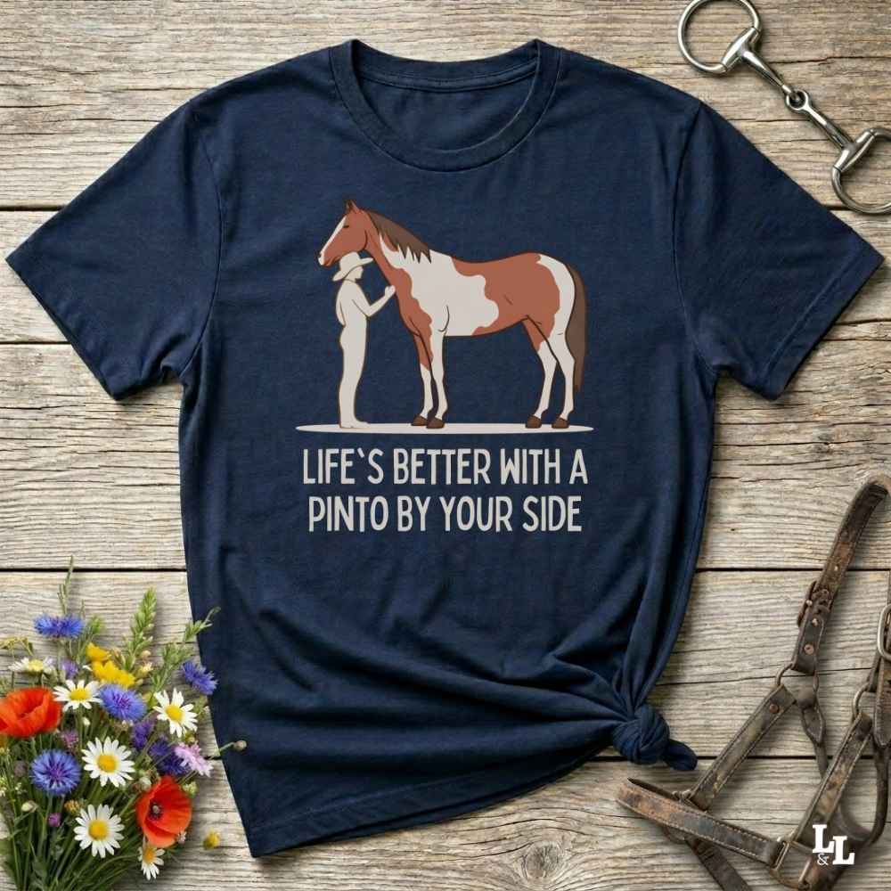 A navy blue graphic t-shirt laid flat on a rustic wooden surface, styled with equestrian accessories and a small bouquet of wildflowers. The shirt features a graphic of a Pinto horse and a person, with the text 'LIFE'S BETTER WITH A PINTO BY YOUR SIDE'.