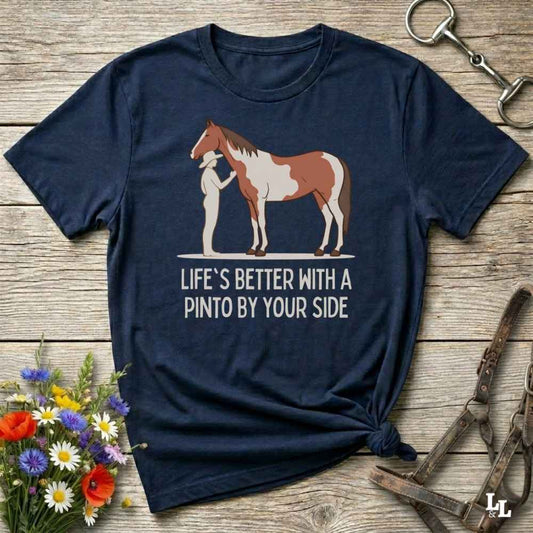 A navy blue graphic t-shirt laid flat on a rustic wooden surface, styled with equestrian accessories and a small bouquet of wildflowers. The shirt features a graphic of a Pinto horse and a person, with the text 'LIFE'S BETTER WITH A PINTO BY YOUR SIDE'.