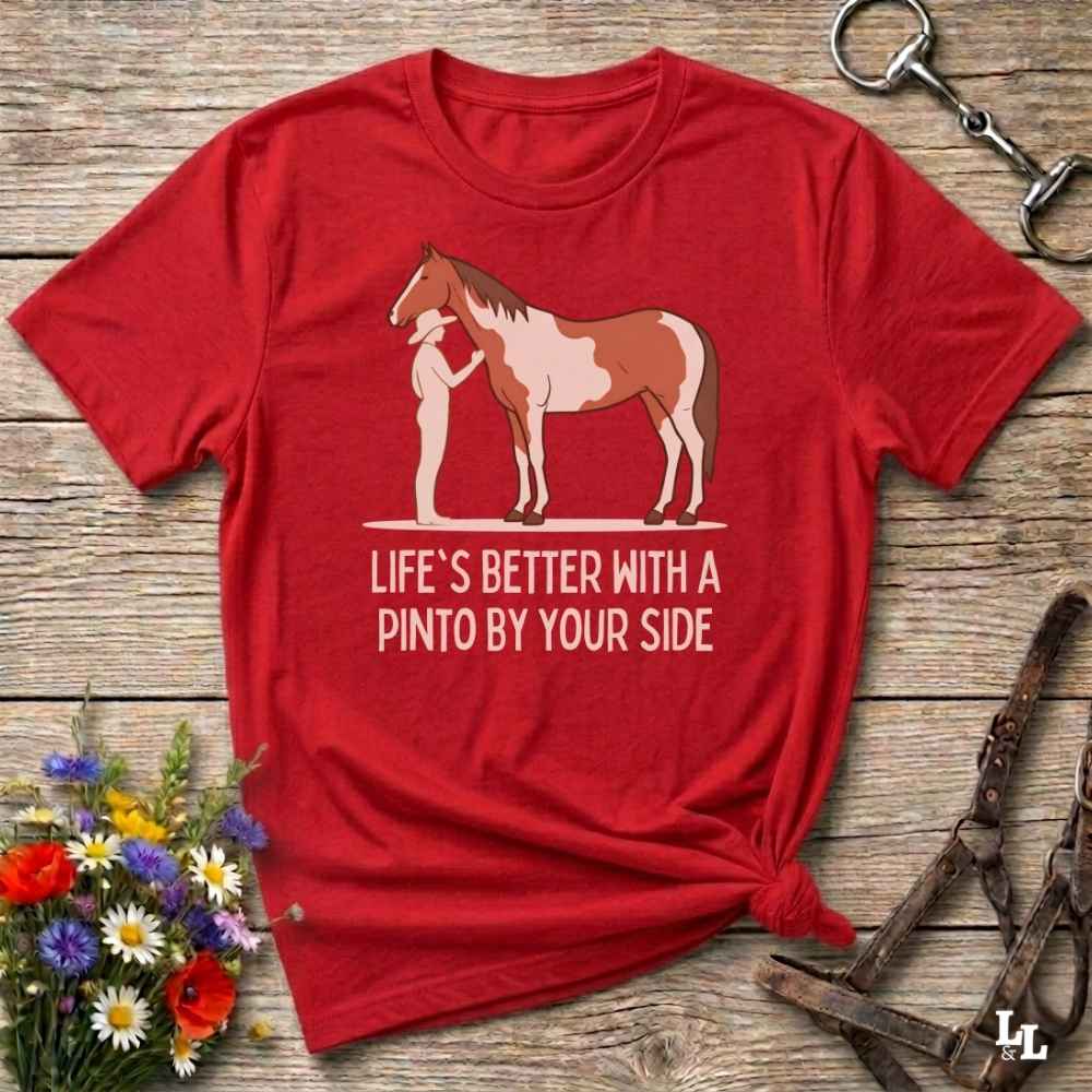A red short-sleeve graphic T-shirt laid flat on a rustic wooden surface, styled with flowers, a horse bit, and a halter. The shirt features an illustration of a pinto horse and a person standing beside it, with the text 'LIFE'S BETTER WITH A PINTO BY YOUR SIDE' printed below.