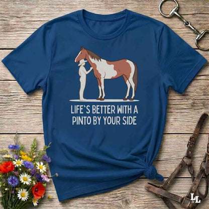 A navy blue graphic t-shirt laid flat on a rustic wooden surface, styled with equestrian accessories and a small bouquet of wildflowers. The shirt features a graphic of a Pinto horse and a person, with the text 'LIFE'S BETTER WITH A PINTO BY YOUR SIDE'.