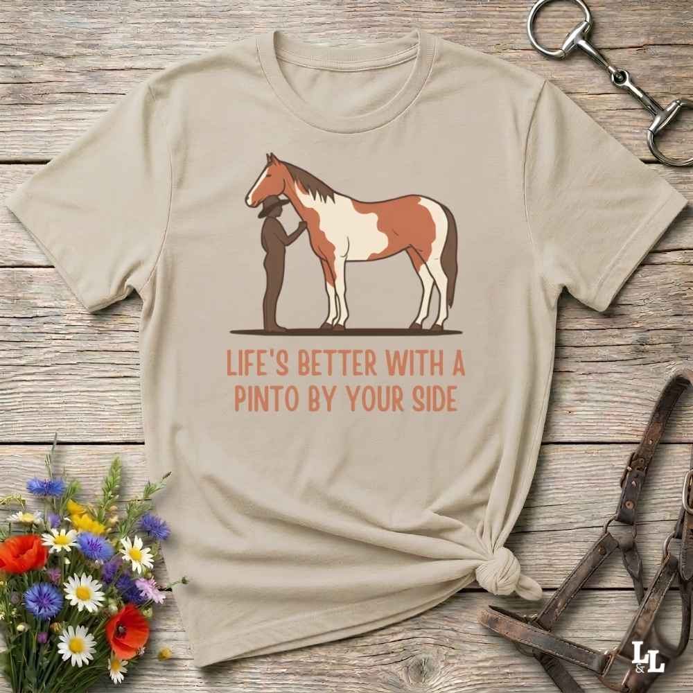 A beige graphic t-shirt featuring a brown and white pinto horse and shadow illustration with the text 'LIFE'S BETTER WITH A PINTO BY YOUR SIDE' in orange. The shirt is styled in a flat-lay on a rustic wooden background with equestrian accessories and a small bouquet of wildflowers.
