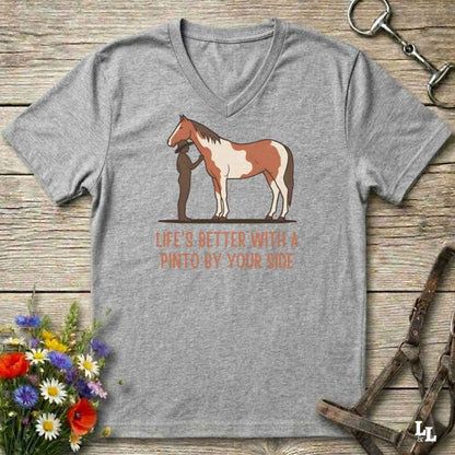 A heather gray V-neck t-shirt laid flat on a rustic wooden surface, featuring a graphic of a Pinto horse and a person with the text 'LIFE'S BETTER WITH A PINTO BY YOUR SIDE' in orange. Decorative flowers and horse tack are placed around the shirt.