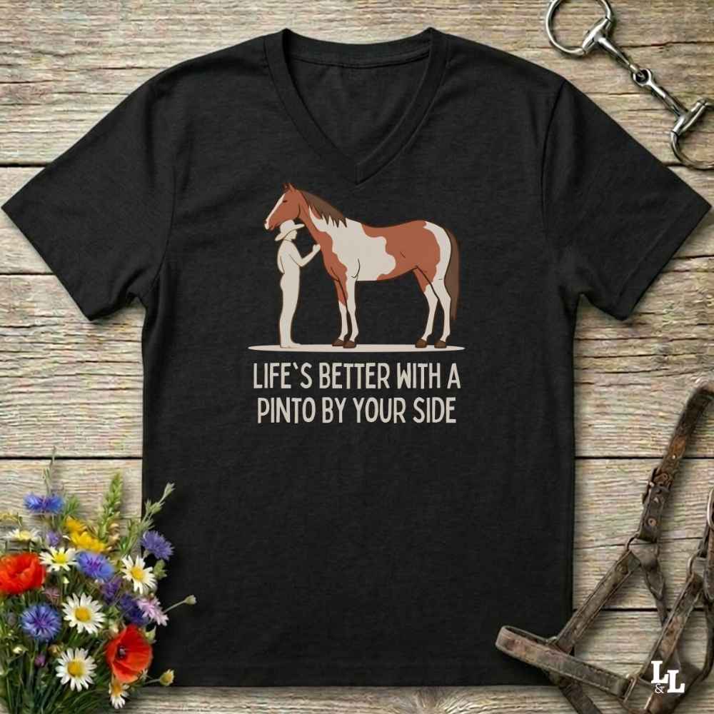 A black V-neck t-shirt laid flat on a rustic wooden surface, featuring a graphic of a brown and white paint horse with a person standing beside it, and the text 'LIFE'S BETTER WITH A PINTO BY YOUR SIDE'. The shirt is styled with equestrian accessories (a halter and bit) and a small bouquet of wildflowers.