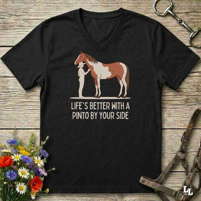A black V-neck t-shirt laid flat on a rustic wooden surface, featuring a graphic of a brown and white paint horse with a person standing beside it, and the text 'LIFE'S BETTER WITH A PINTO BY YOUR SIDE'. The shirt is styled with equestrian accessories (a halter and bit) and a small bouquet of wildflowers.
