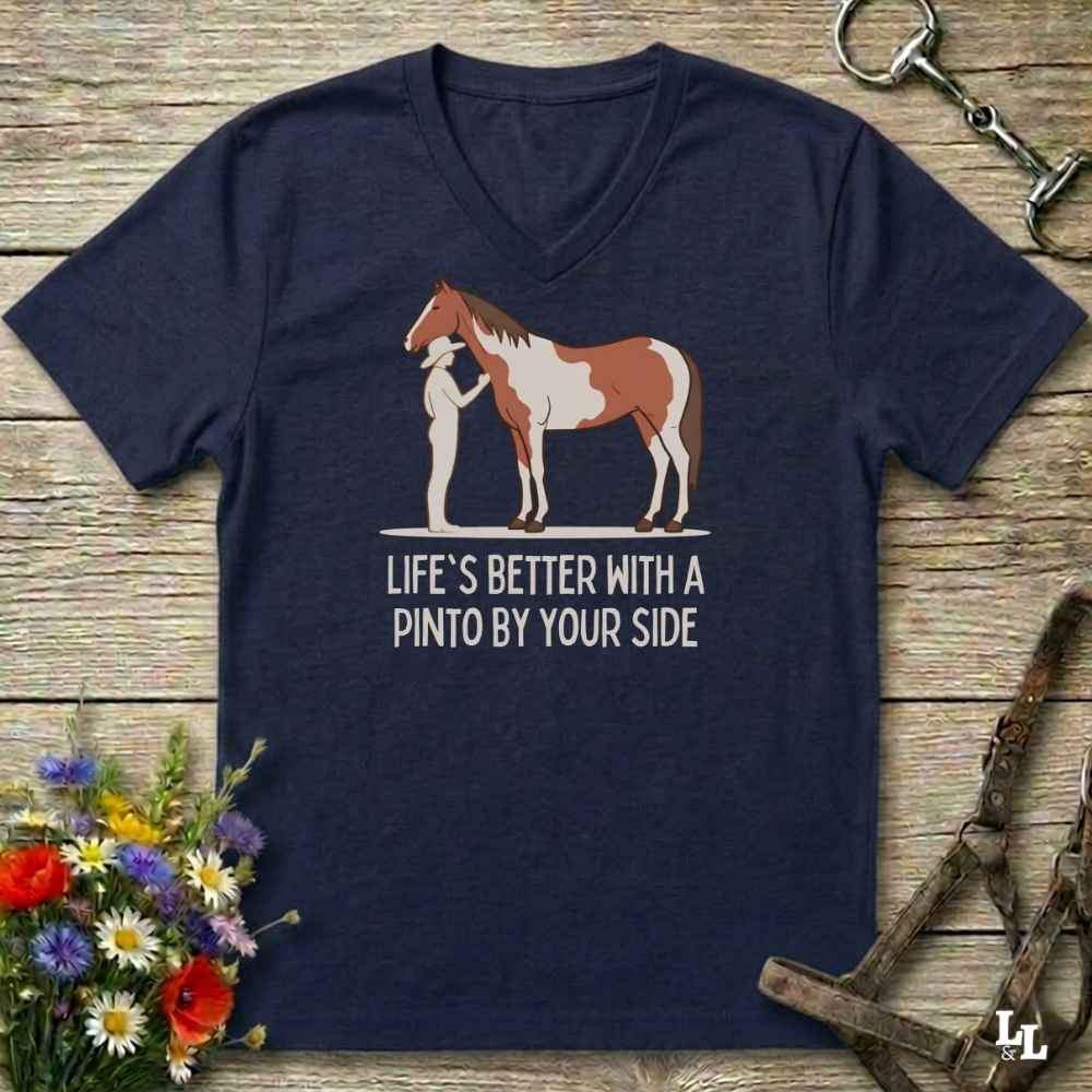 A navy blue V-neck t-shirt laid flat on a rustic wooden surface, featuring a graphic of a Pinto horse and a person with the text 'LIFE'S BETTER WITH A PINTO BY YOUR SIDE'. The shirt is styled with equestrian accessories (bridle, halter, tack) and a small bouquet of wildflowers.