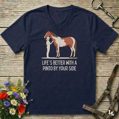 A navy blue V-neck t-shirt laid flat on a rustic wooden surface, featuring a graphic of a Pinto horse and a person with the text 'LIFE'S BETTER WITH A PINTO BY YOUR SIDE'. The shirt is styled with equestrian accessories (bridle, halter, tack) and a small bouquet of wildflowers.
