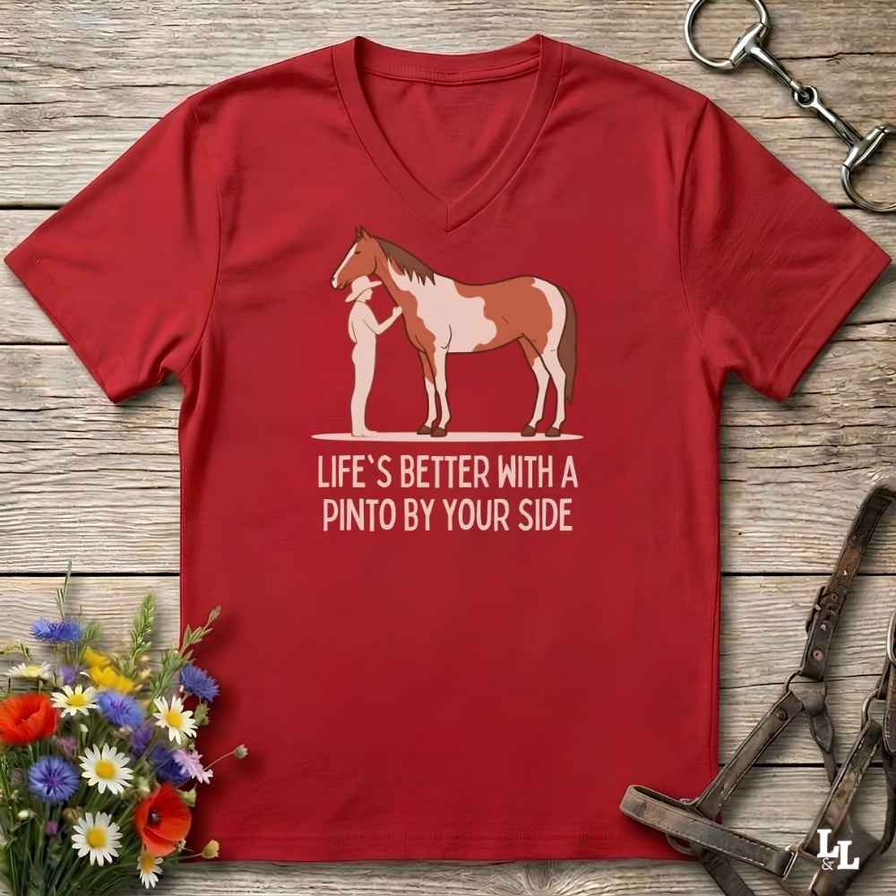 A red V-neck t-shirt laid flat on a rustic wood surface, featuring a graphic of a white horse with brown patches standing next to a white figure, with the text 'LIFE'S BETTER WITH A PINTO BY YOUR SIDE' printed below. The shirt is styled with equestrian accessories and a small bouquet of wildflowers.