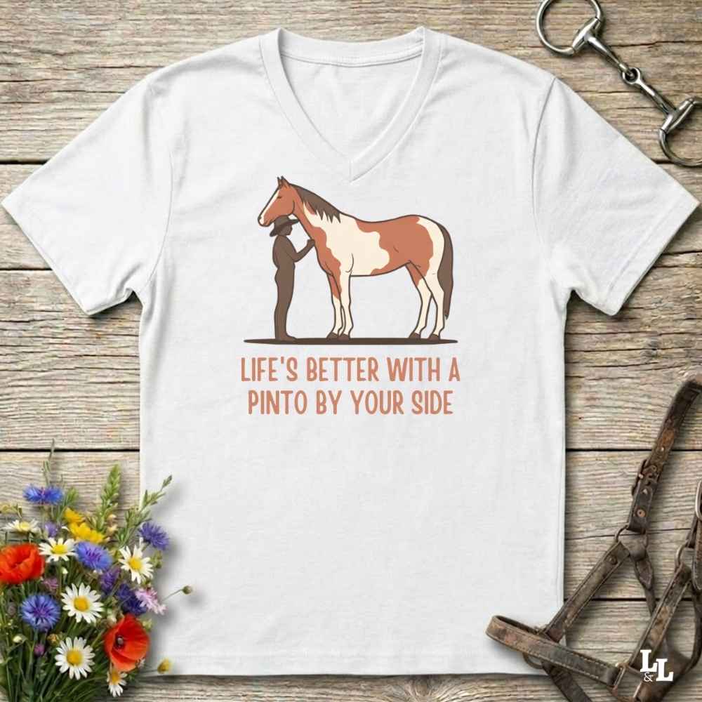 A white V-neck t-shirt laid flat on a rustic wooden surface, featuring a graphic of a Pinto horse and a person with the text 'LIFE'S BETTER WITH A PINTO BY YOUR SIDE' in orange. The shirt is styled with horse tack, flowers, and a leather strap.
