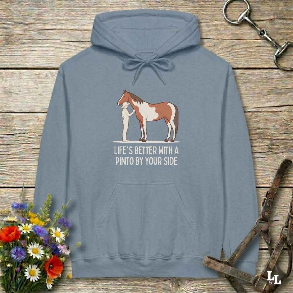 A slate blue pullover hoodie laid flat on a rustic wooden surface, featuring a graphic of a Pinto horse and a person with the text 'LIFE'S BETTER WITH A PINTO BY YOUR SIDE'. The hoodie is styled with equestrian props including a halter, tack, and a small bouquet of wildflowers.