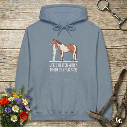 A slate blue pullover hoodie laid flat on a rustic wooden surface, featuring a graphic of a Pinto horse and a person with the text 'LIFE'S BETTER WITH A PINTO BY YOUR SIDE'. The hoodie is styled with equestrian props including a halter, tack, and a small bouquet of wildflowers.