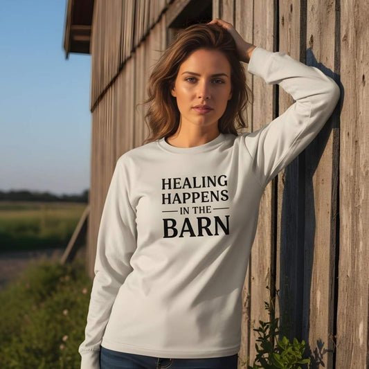 Woman wearing a white sweatshirt with 'Healing Happens in the Barn' text, standing in front of a wooden barn. Healing Happens in the Barn Long Sleeve Tee for Horse Lovers, Trail Riding Tee