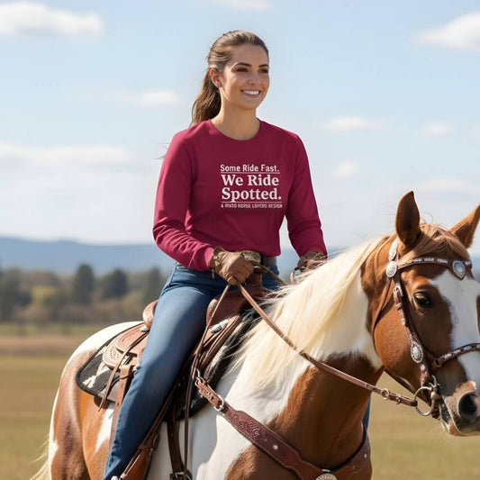 Woman riding a horse wearing a maroon sweatshirt with text, set against a scenic background. We Ride Spotted Long Sleeve Tee for Pinto Horse Lovers, Trail Riding Tee