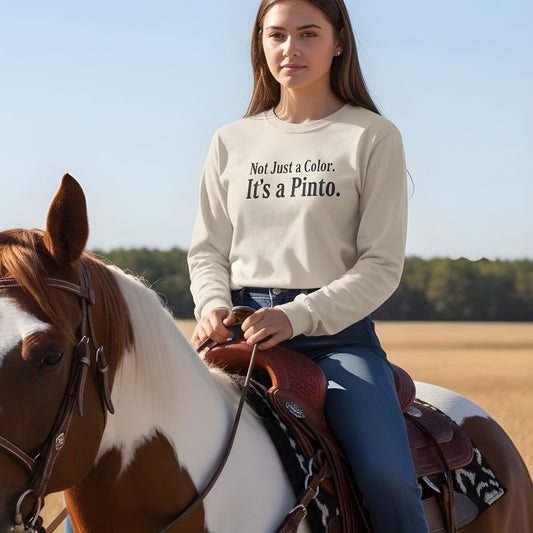 Woman riding a horse wearing a sweatshirt with text, set against a clear sky and open field. Not Just a Color Long Sleeve Tee for Pinto Lovers, Trail Riding Tee
