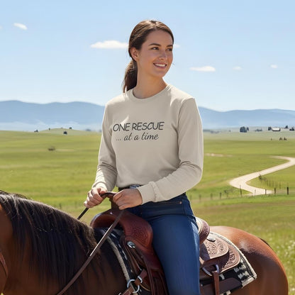 Woman riding a horse in a field with mountains in the background, wearing a shirt with text. One Rescue at a Time Long Sleeve Tee for Horse Lovers, Trail Riding Tee