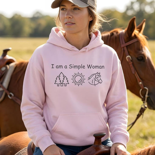Woman wearing a pink hoodie with nature-themed design and text, sitting on a horse in a field. I am a Simple Woman Hoodie for Horse Lovers, Equestrian Apparel, Trail Riding Hoodie