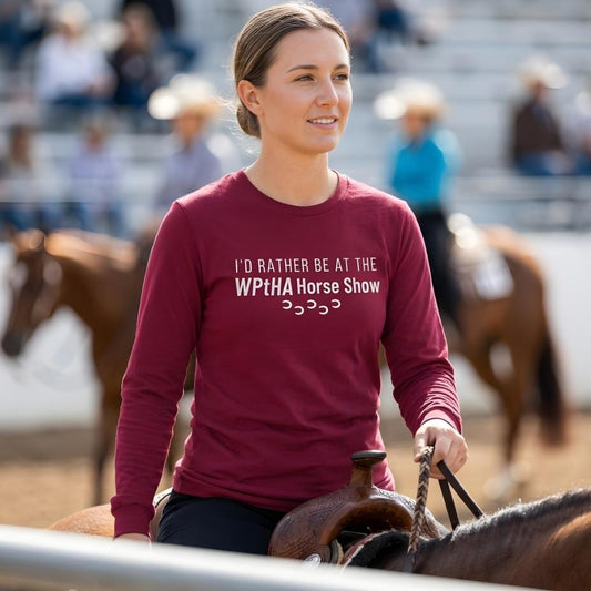Woman in a red shirt with horse show text, standing next to a horse in an outdoor setting.  I'd Rather be at the WPtHA Horse Show Long Sleeve Tee for Pinto Horse Lovers. Charity tee raising funds for Wisconsin Pinto Horse Association