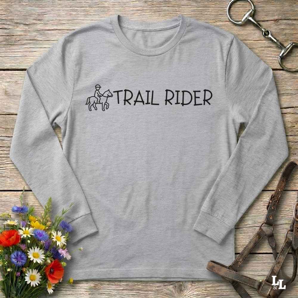A light gray long-sleeve t-shirt with a black graphic of an equestrian rider and the words 'TRAIL RIDER' printed on the chest, laid flat on a rustic wooden surface with equestrian accessories and flowers as props.