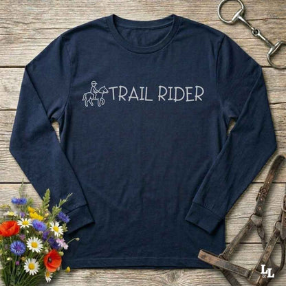 A navy blue long-sleeve t-shirt with a white graphic reading 'TRAIL RIDER' and a simple rider-and-horse outline, laid flat on a rustic wood surface with flowers, horse tack, and a bridle around it.