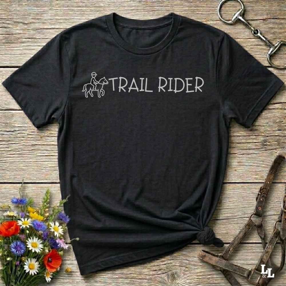 Black t-shirt with 'Trail Rider' text and graphic on a wooden surface with floral and equestrian equipment.