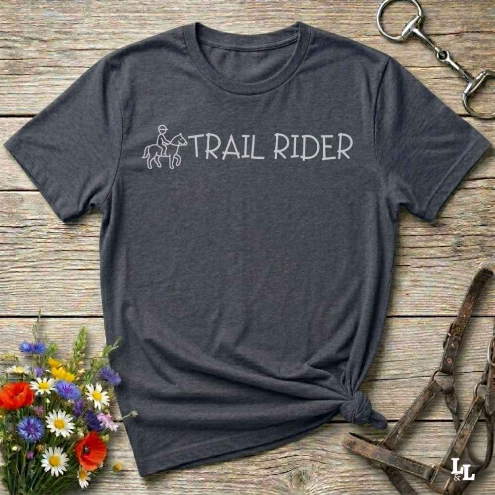 A dark heather gray t-shirt with a white graphic reading 'TRAIL RIDER' and a small rider-and-horse outline, laid flat on a rustic wood surface with flowers and equestrian gear around it.