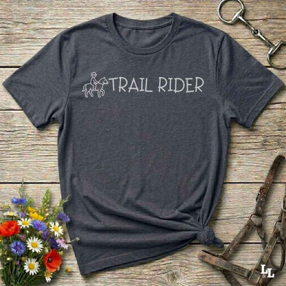 A dark heather gray t-shirt with a white graphic reading 'TRAIL RIDER' and a small rider-and-horse outline, laid flat on a rustic wood surface with flowers and equestrian gear around it.
