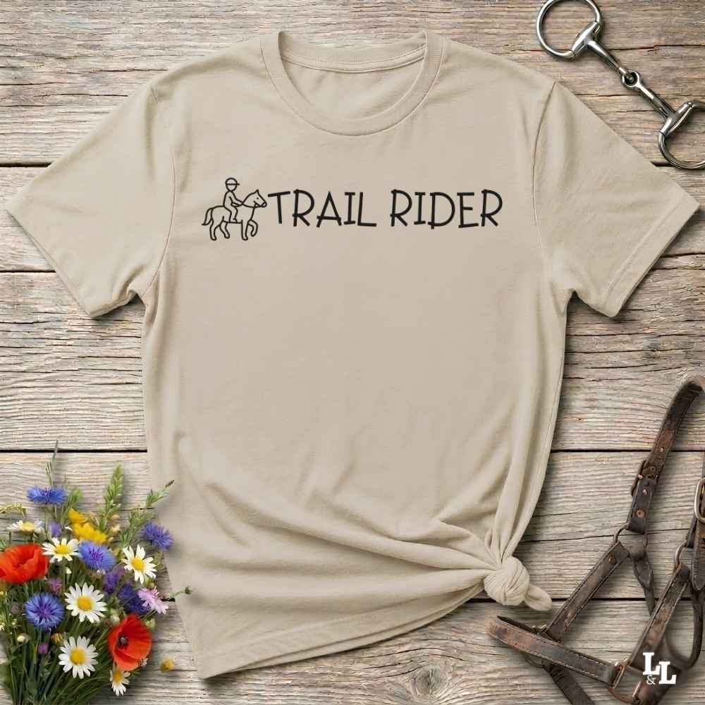 A beige short-sleeve T-shirt laid flat on a rustic wood surface, with a black 'TRAIL RIDER' graphic and small rider-and-horse illustration across the chest. The shirt is styled with flowers, a horse bit, and a leather bridle around it.