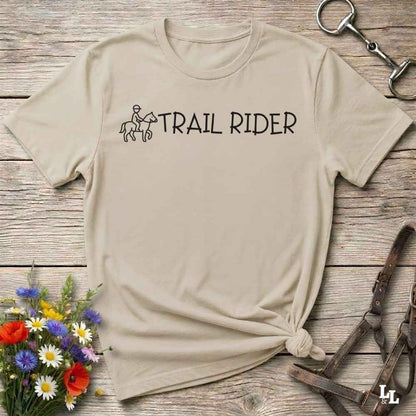 A beige short-sleeve T-shirt laid flat on a rustic wood surface, with a black 'TRAIL RIDER' graphic and small rider-and-horse illustration across the chest. The shirt is styled with flowers, a horse bit, and a leather bridle around it.
