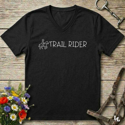 A black  V-neck t-shirt with a black graphic reading 'TRAIL RIDER' and a small rider-and-horse illustration, laid flat on a rustic wood surface with flowers and equestrian accessories 