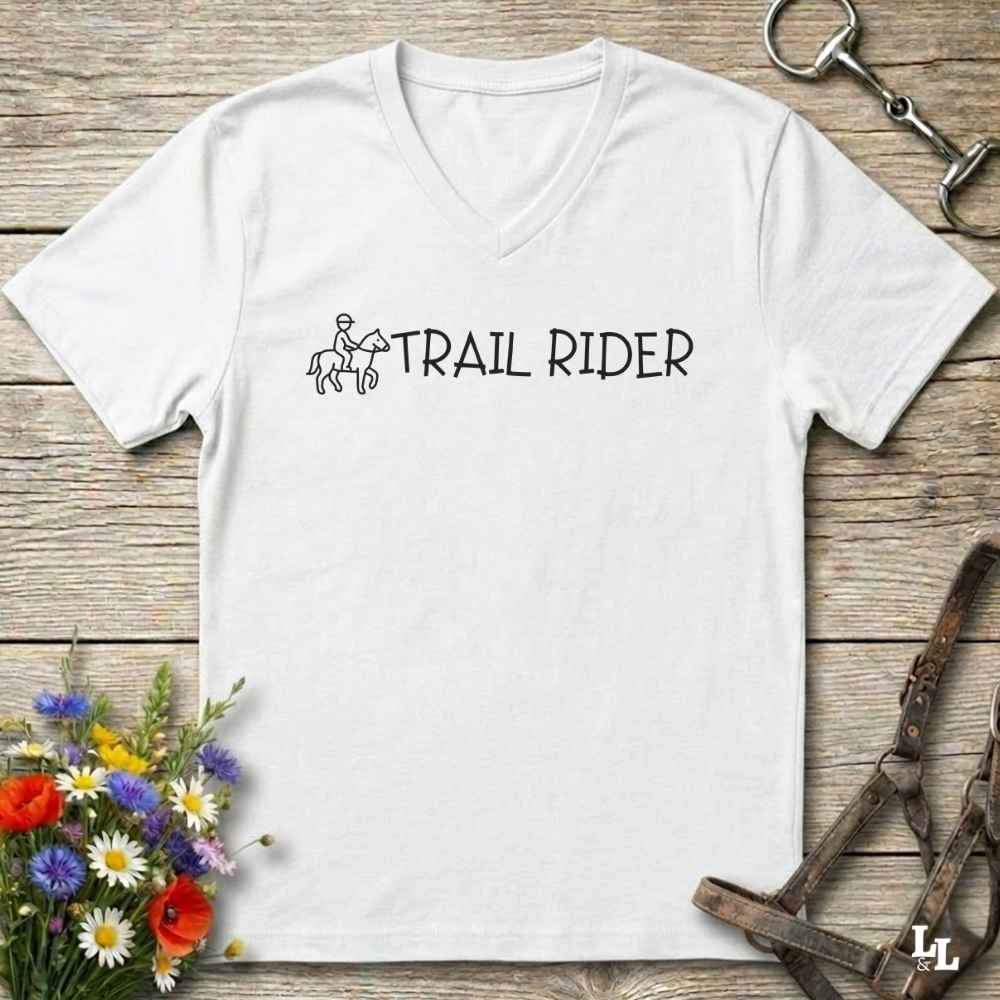 A white V-neck t-shirt with a black graphic reading 'TRAIL RIDER' and a small rider-and-horse illustration, laid flat on a rustic wood surface with flowers and equestrian accessories 