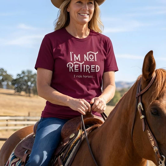 Woman riding a horse wearing a maroon t-shirt with text, with a clear blue sky and trees in the background. Beige I'm Not Retired, I Ride Horses for Horse Lovers, Trail Riding Tee, Equestrian Apparel, great gift for horse person