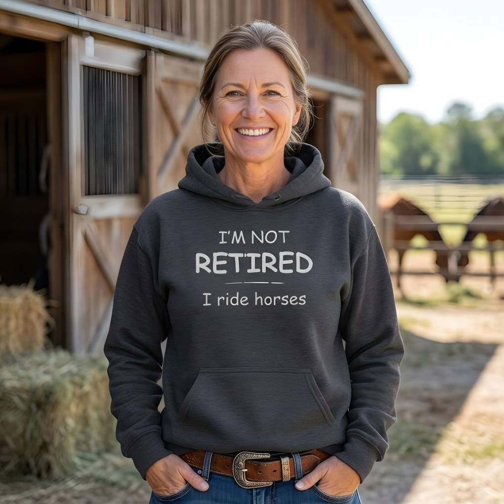 Woman wearing a hoodie with 'I'm not retired, I ride horses' text in front of a barn with horses.  I'm Not Retired I Ride Horses Hoodie for Horse Riders, Equestrian Cozy Hoodie, perfect gift for horse lovers