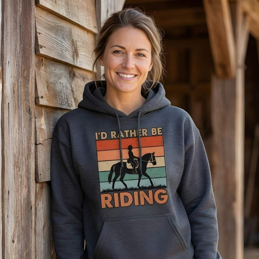 Woman wearing a hoodie with 'I'd Rather Be Riding' graphic leaning against a wooden building. I'd Rather be Riding Hoodie for Horse Riders, Cozy Barn Wear Hoodie, perfect gift for horse lovers