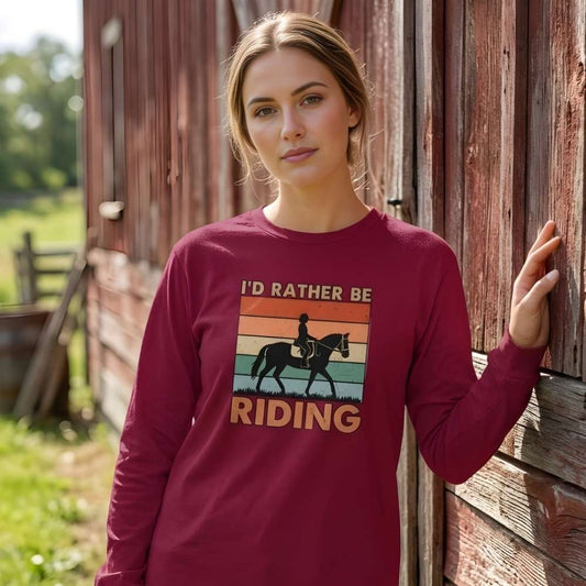 Woman wearing a maroon long-sleeve shirt with a horse graphic and text, standing next to a barn. I'd Rather be Riding Longsleeve T-shirt for Horse Riders, Trail Riding Tee, perfect gift for horse lovers