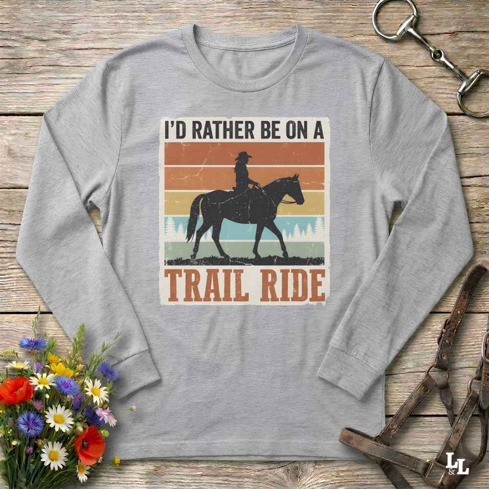 A light gray long-sleeve t-shirt with a retro-style graphic of a cowboy riding a horse, surrounded by the text 'I'D RATHER BE ON A TRAIL RIDE'. The shirt is displayed flat on a rustic wooden background with decorative flowers and leather tack.