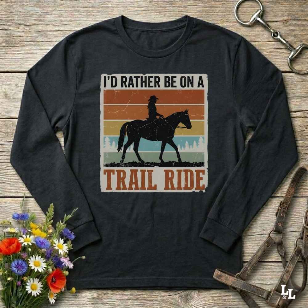 A black long-sleeve t-shirt with a vintage-style western graphic that reads 'I'D RATHER BE ON A TRAIL RIDE' with a silhouette of a cowboy on a horse, displayed on a rustic wooden background with decorative flowers and horse tack.