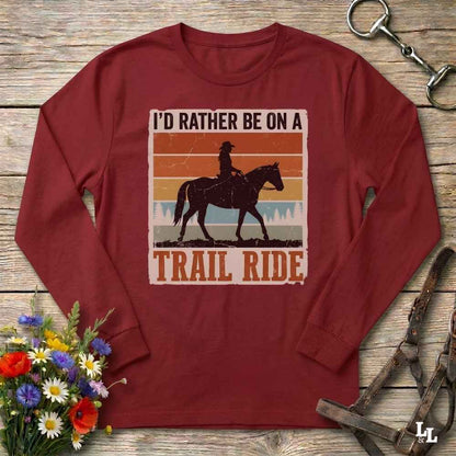 A long-sleeve maroon crewneck t-shirt with a retro-style graphic of a trail rider on a horse, surrounded by the text 'I'D RATHER BE ON A TRAIL RIDE'. The shirt is displayed flat on a rustic wooden background with decorative props including a bouquet of flowers, a horse bit, and a leather bridle.