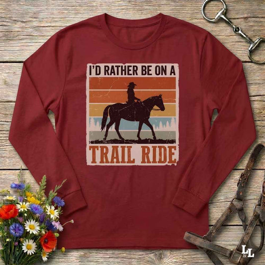 A long-sleeve maroon crewneck t-shirt with a retro-style graphic of a trail rider on a horse, surrounded by the text 'I'D RATHER BE ON A TRAIL RIDE'. The shirt is displayed flat on a rustic wooden background with decorative props including a bouquet of flowers, a horse bit, and a leather bridle.