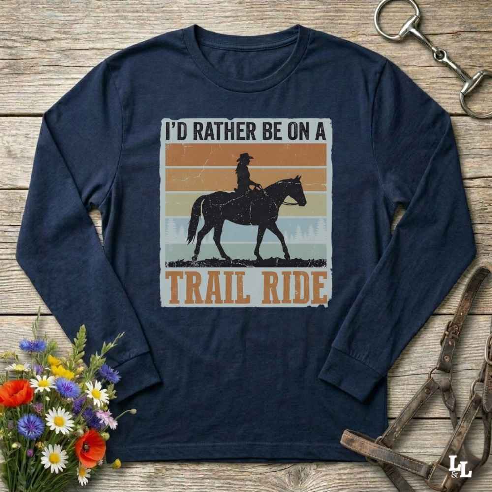 A navy blue long-sleeve t-shirt with a retro-style graphic of a cowboy riding a horse and the text 'I'D RATHER BE ON A TRAIL RIDE'. The shirt is displayed flat on a rustic wooden background with decorative equestrian and floral props.