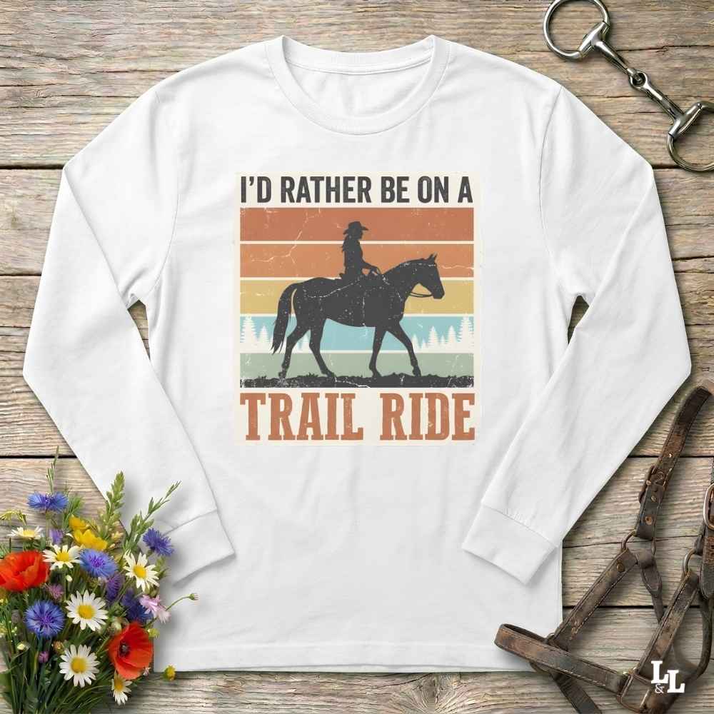 A white long-sleeve t-shirt with a retro-style graphic print of a cowboy riding a horse, accompanied by the text 'I'D RATHER BE ON A TRAIL RIDE'. The shirt is displayed flat on a rustic wooden background with equestrian accessories and flowers.