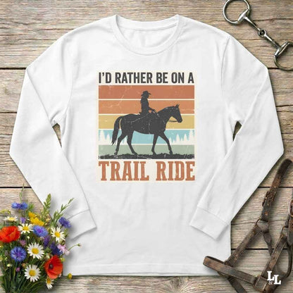 A white long-sleeve t-shirt with a retro-style graphic print of a cowboy riding a horse, accompanied by the text 'I'D RATHER BE ON A TRAIL RIDE'. The shirt is displayed flat on a rustic wooden background with equestrian accessories and flowers.